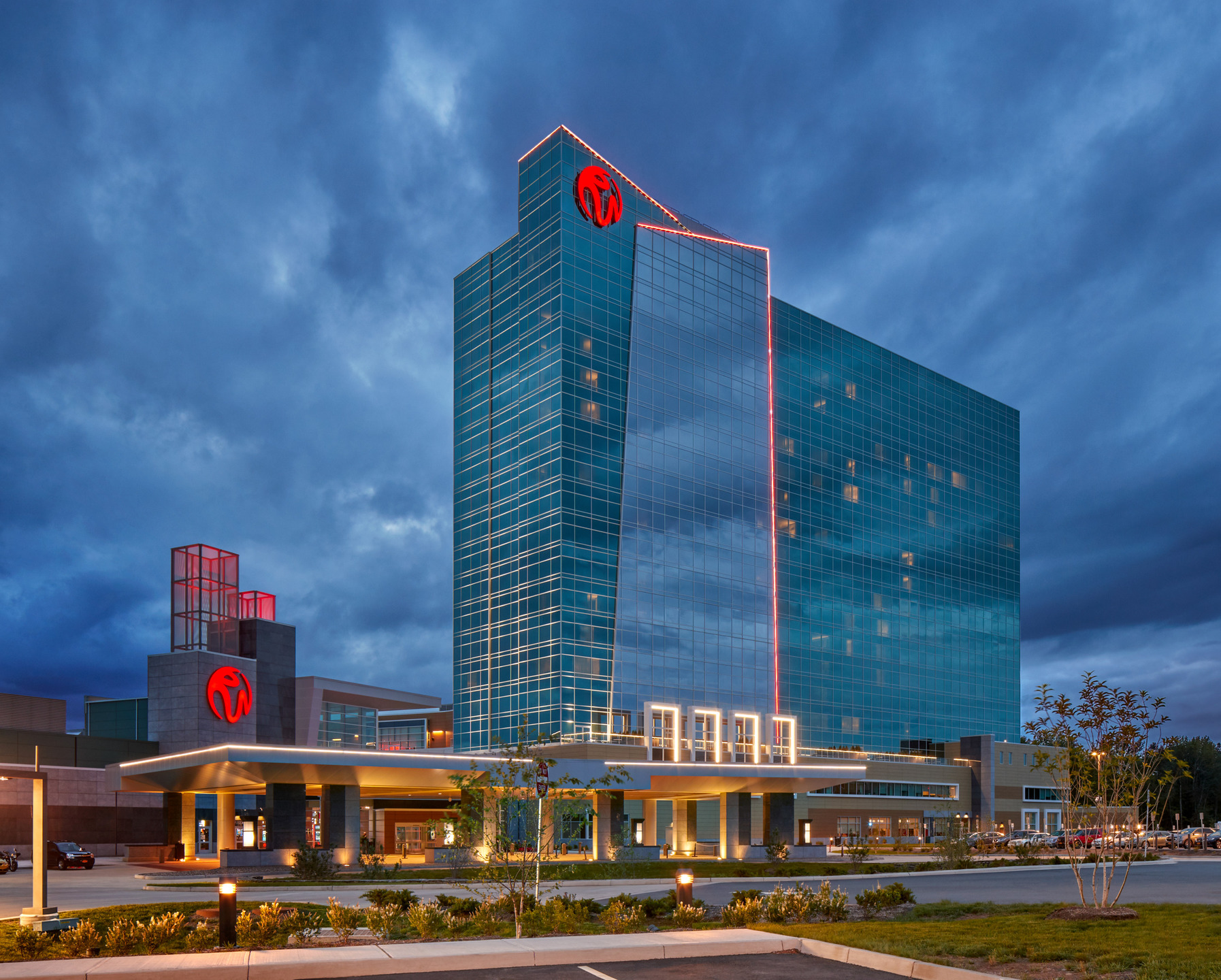 Exterior of a glass tower and entrance at dusk, with illuminated red accents.