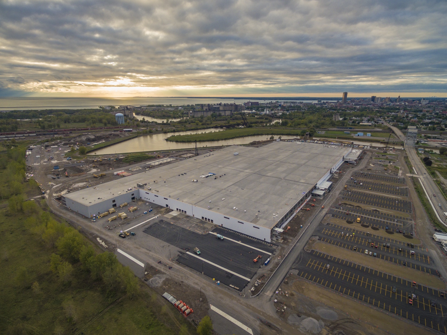 Aerial view of a large construction project beside a river, with the city skyline beyond.